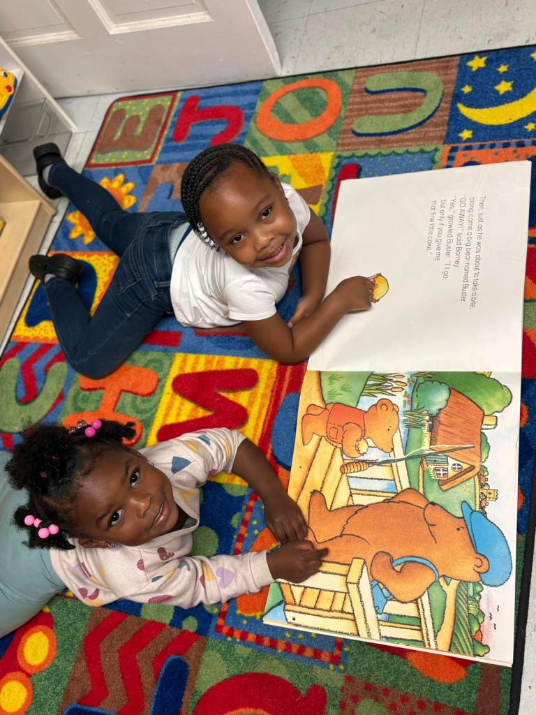 Two children smiling and reading a picture book together on a colorful classroom rug